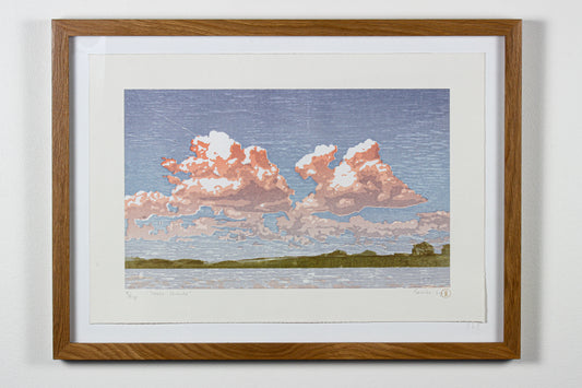 Framed artwork of a landscape with pink clouds over water on a white wall.