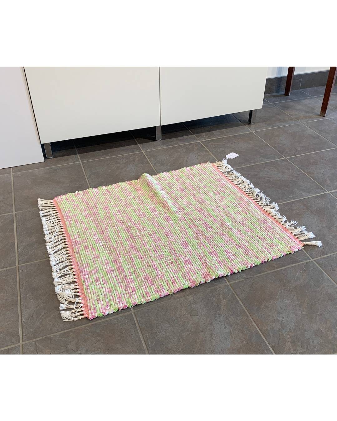 Pink and Green woven rug with fringe laying in front of a cabinet
