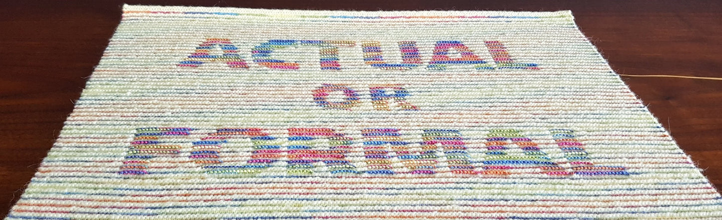 Angle two.; in this angle, the text is legible  Knitted wall hanging in cream and rainbow gradient yarn. Text: ACTUAL OR FORMAL