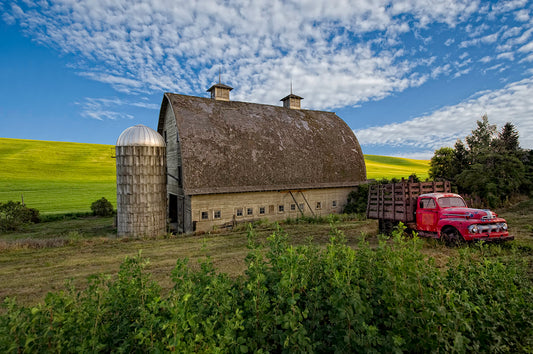 Photograph of a brown barn against yellow and green fields with a red truck