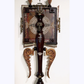 An ornate wall-mounted decorative sculpture featuring a dark reddish-brown central column, carved wing-like side supports, a rectangular panel with a circular lace-like motif, and brass-toned hardware, shown against a plain light wall.