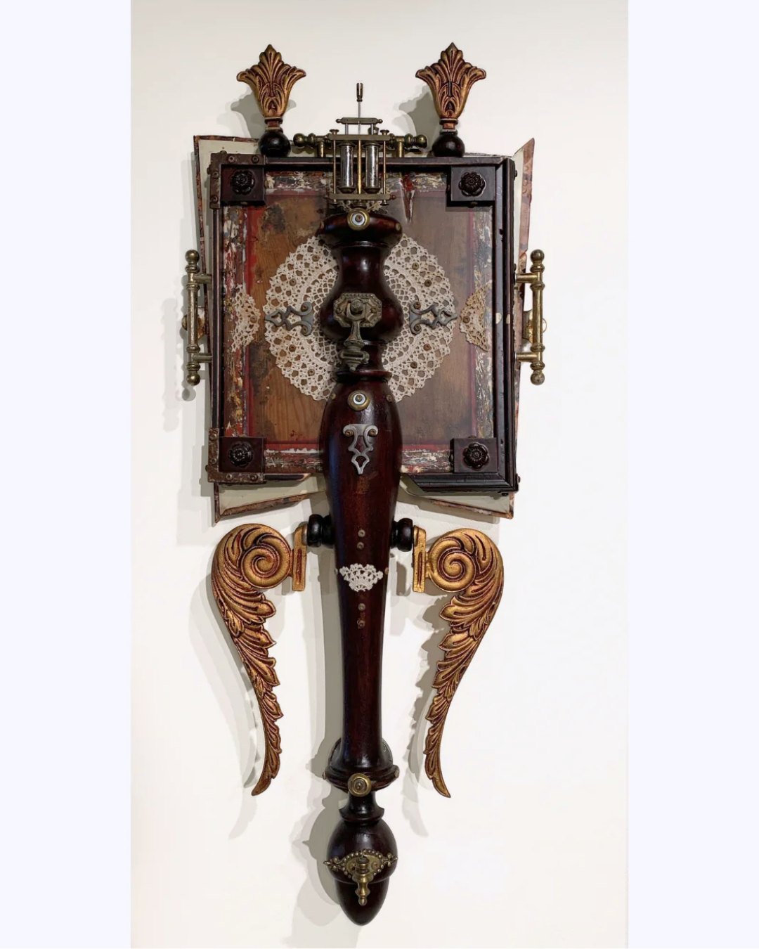 An ornate wall-mounted decorative sculpture featuring a dark reddish-brown central column, carved wing-like side supports, a rectangular panel with a circular lace-like motif, and brass-toned hardware, shown against a plain light wall.