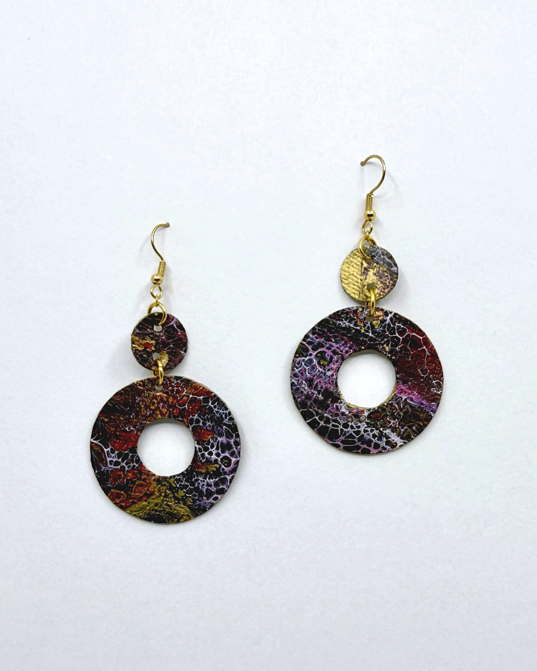 A pair of drop earrings with large circular disc charms featuring a marbled pattern of purple, red, black, and gold, suspended from gold hooks with a small textured gold disc in between.