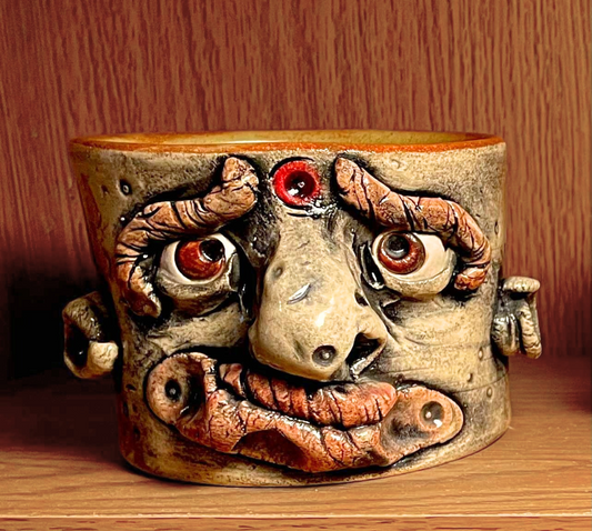 A handmade ceramic mug shaped like a stylized, expressive face with raised eyes, a large nose, and a wide mouth, sitting on a wooden shelf against a wood-panelled background.