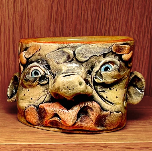 A handmade ceramic mug shaped like a stylized face with wide blue eyes, textured brown and beige tones, and reddish-orange highlights, set against a wooden background.