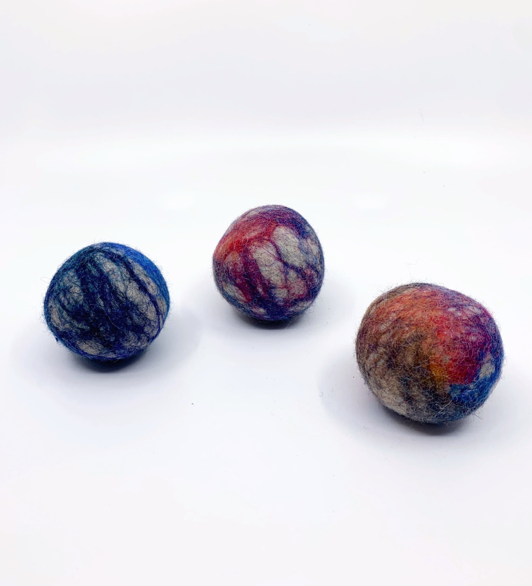Three hand-felted wool balls in marbled colors of blue, purple, red, and white, arranged on a plain white background.