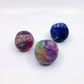 Three hand-felted wool balls in marbled colors, including blue, purple, pink, and green, arranged on a plain white background.