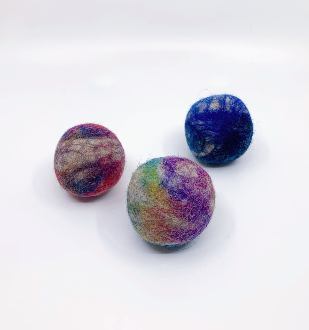 Three hand-felted wool balls in marbled colors, including blue, purple, pink, and green, arranged on a plain white background.