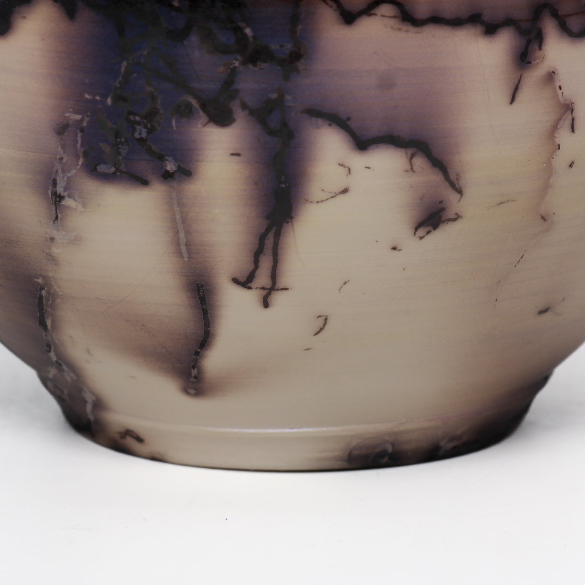 A round ceramic vessel. Surface features black and brown lines from burnt horsehair, with dark brown splotches on a beige surface.