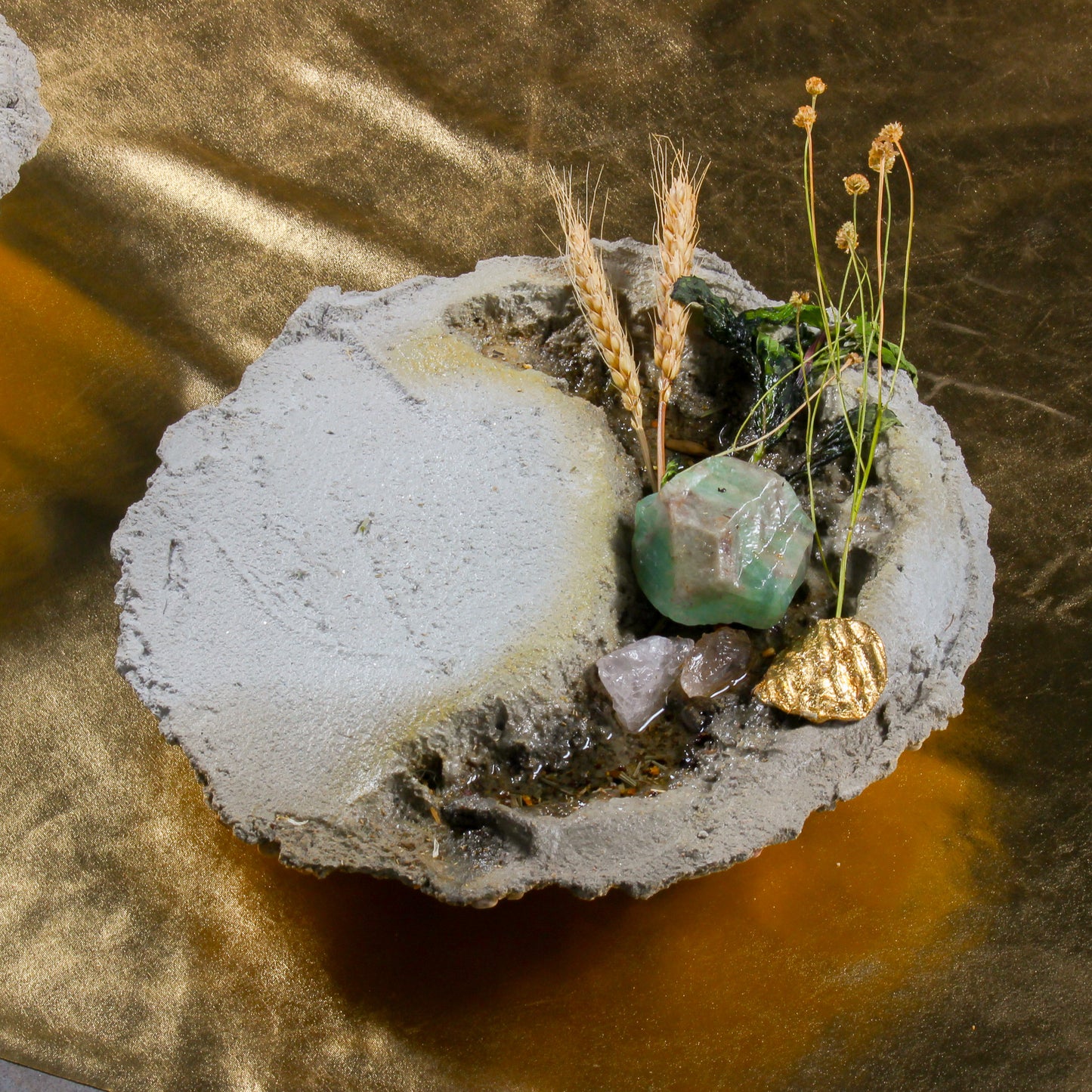 A close up shot of one of the bowl shaped objects. It is made of cement, with pieces of plant life, crystals, a gold chip, and epoxy that looks like water sitting inside.