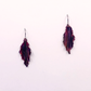 A pair of handmade dangle earrings with a marbled pattern of purple, copper, and black, suspended from silver fishhook wires against a plain white background.