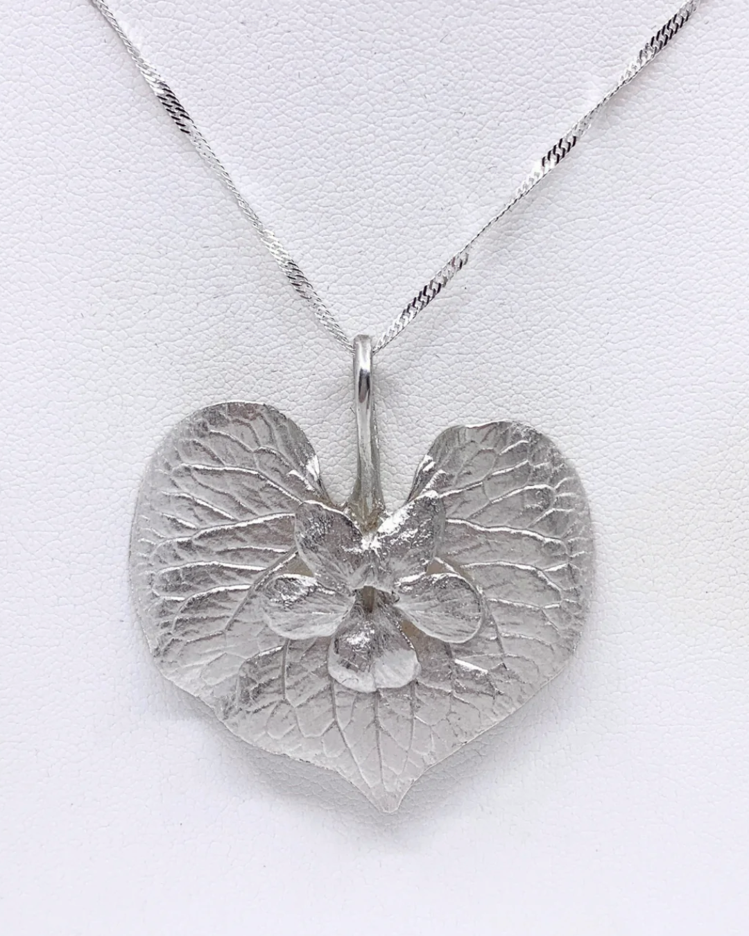 A silver heart-shaped pendant necklace with a textured, leaf-like surface and a small central floral detail, suspended from a twisted silver chain against a white background.