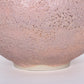 Close up shot of a round ceramic vessel. The base is smooth and symmetrical, with pink textured glaze on the outside.