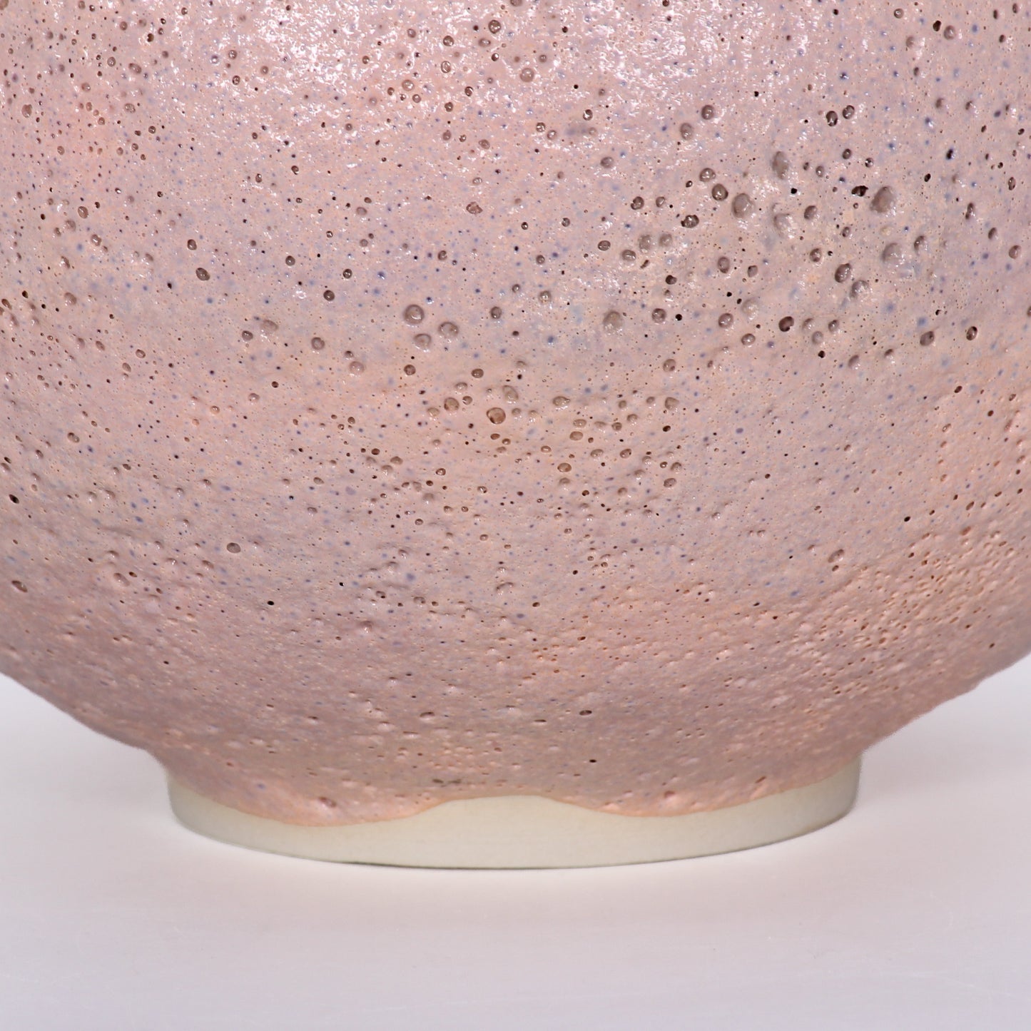 Close up shot of a round ceramic vessel. The base is smooth and symmetrical, with pink textured glaze on the outside.