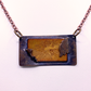 A rectangular pendant necklace featuring a textured dark bronze metal frame with a central gold-toned floral design, suspended from a rose gold-toned chain against a light background.