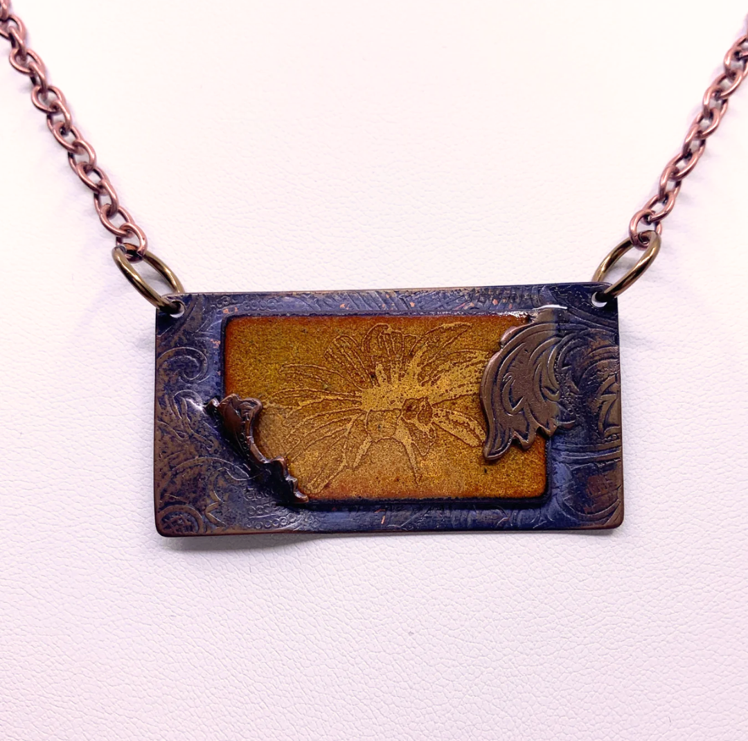 A rectangular pendant necklace featuring a textured dark bronze metal frame with a central gold-toned floral design, suspended from a rose gold-toned chain against a light background.