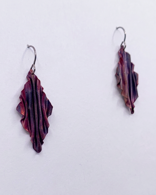 A pair of handmade dangle earrings with textured, layered dark purple and copper pieces hanging from silver fishhook wires, displayed against a plain white background.