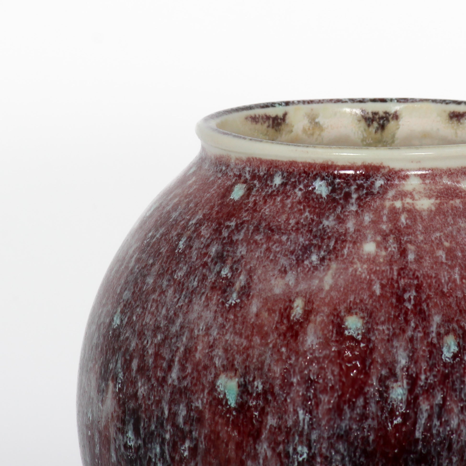 Close up shot of a round ceramic vessel. The mouth is smooth and symmetrical, with spots of burgundy and pale blue textured glaze.