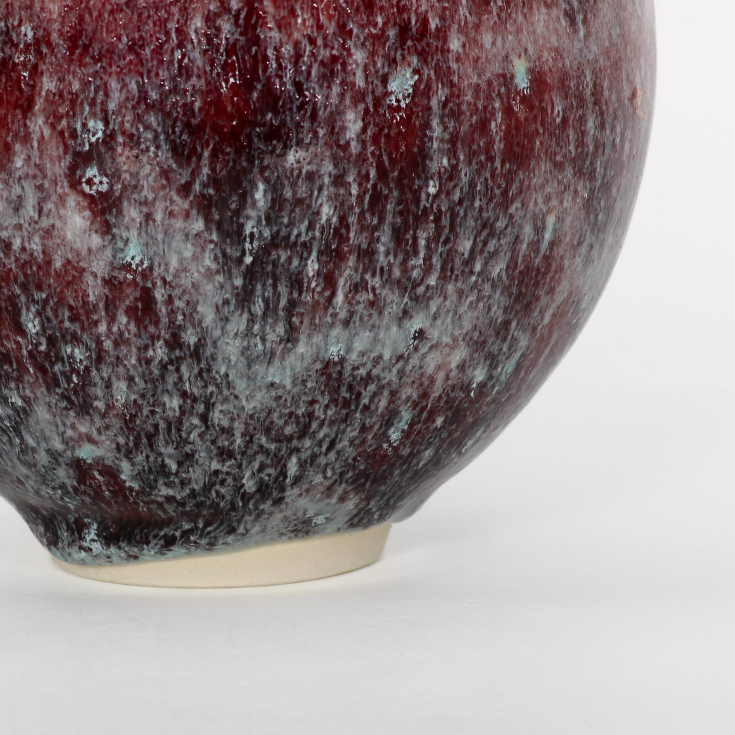 Close up shot of a round ceramic vessel. The base is smooth and symmetrical, with spots of burgundy and pale blue textured glaze.