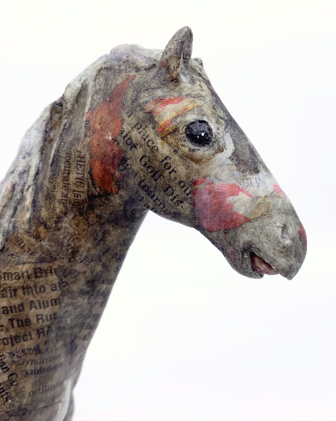 A close-up of a sculpted horse head made from recycled newspaper, featuring printed text and red painted accents, set against a plain white background.