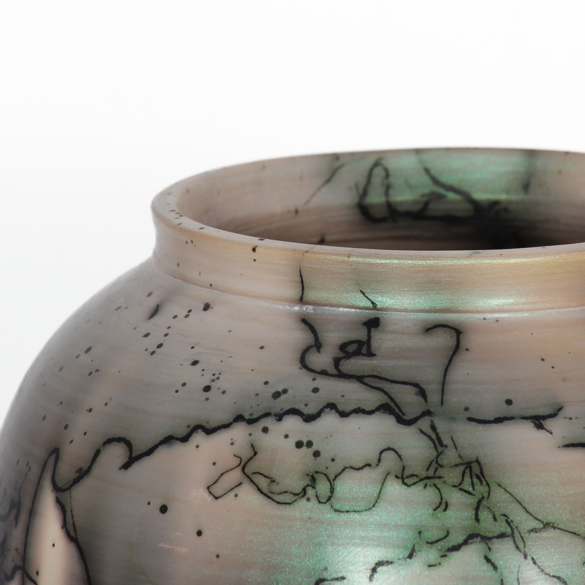 A round ceramic vessel. The clay is beige, with black lines from burnt horsehair, and green and grey spots of glaze. Green spots have a shimmery sheen.