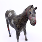 A small horse figurine standing on a plain white background. The horse has a mottled gray and white body with red markings on its face and legs, and a textured, collage-like painted finish.