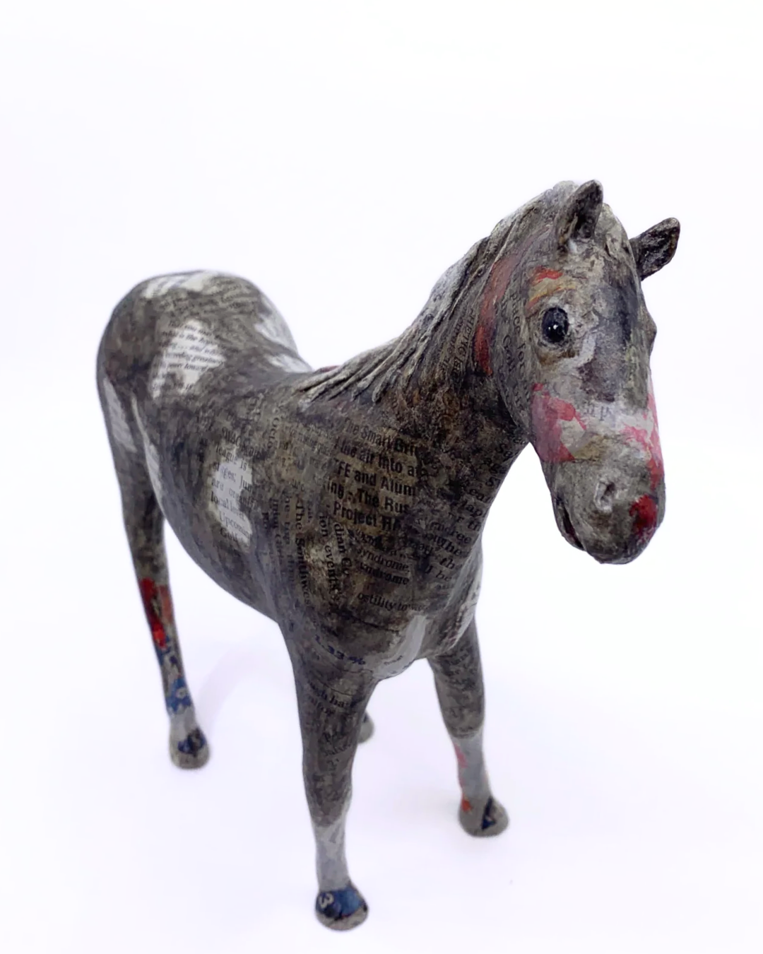 A small horse figurine standing on a plain white background. The horse has a mottled gray and white body with red markings on its face and legs, and a textured, collage-like painted finish.