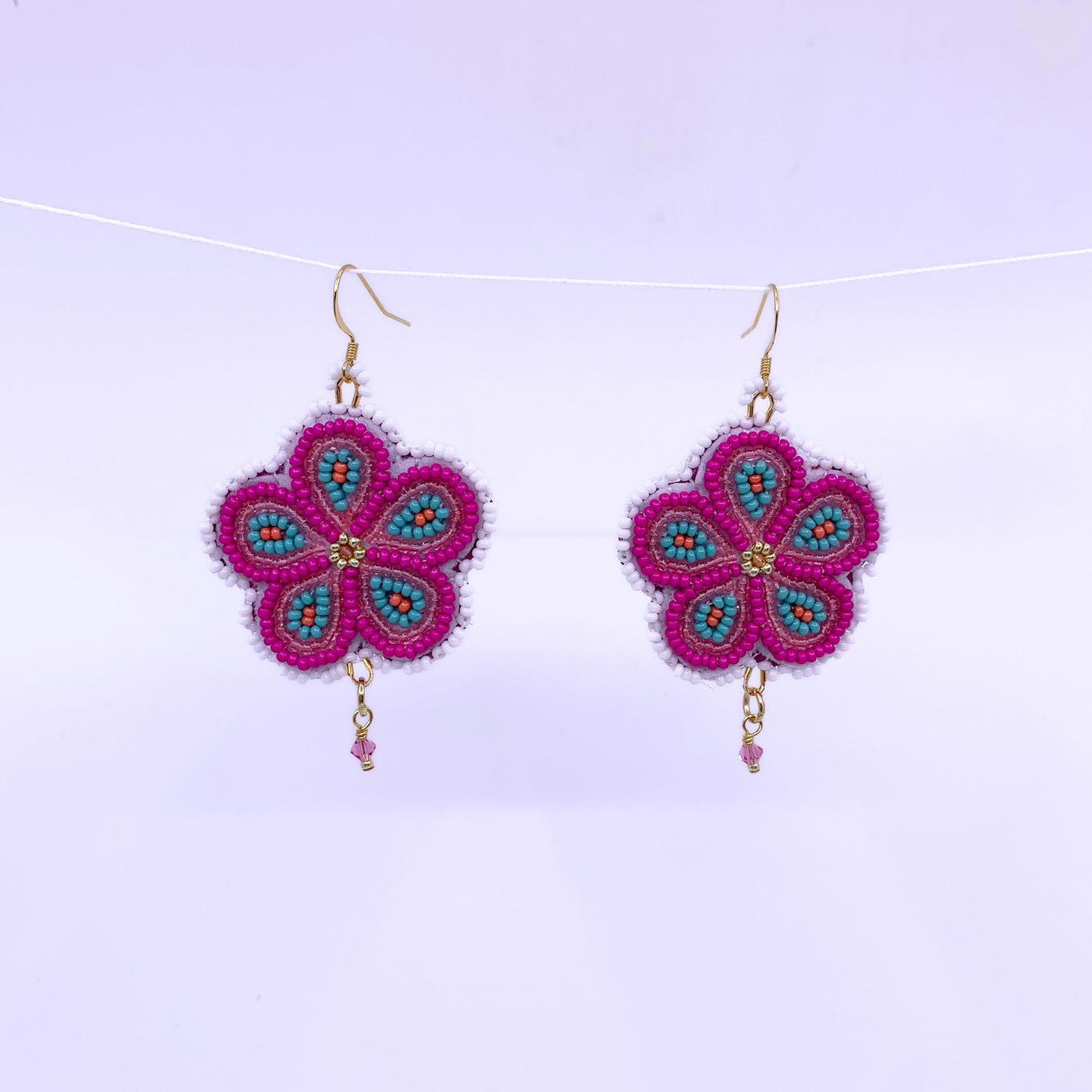 A pair of handmade beaded flower dangle earrings with bright pink petals, turquoise blue accents, and gold hooks, suspended against a light lavender background.