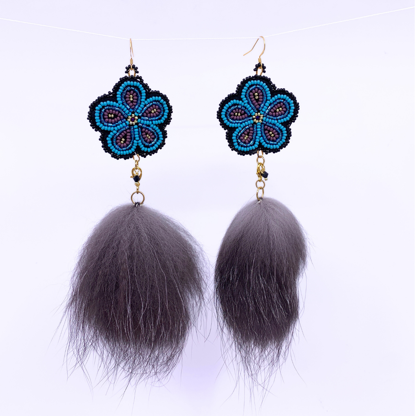 A pair of handmade dangle earrings featuring blue and black beaded flower charms with gold hooks and long, fluffy dark gray faux fur tassels, shown against a plain white background.