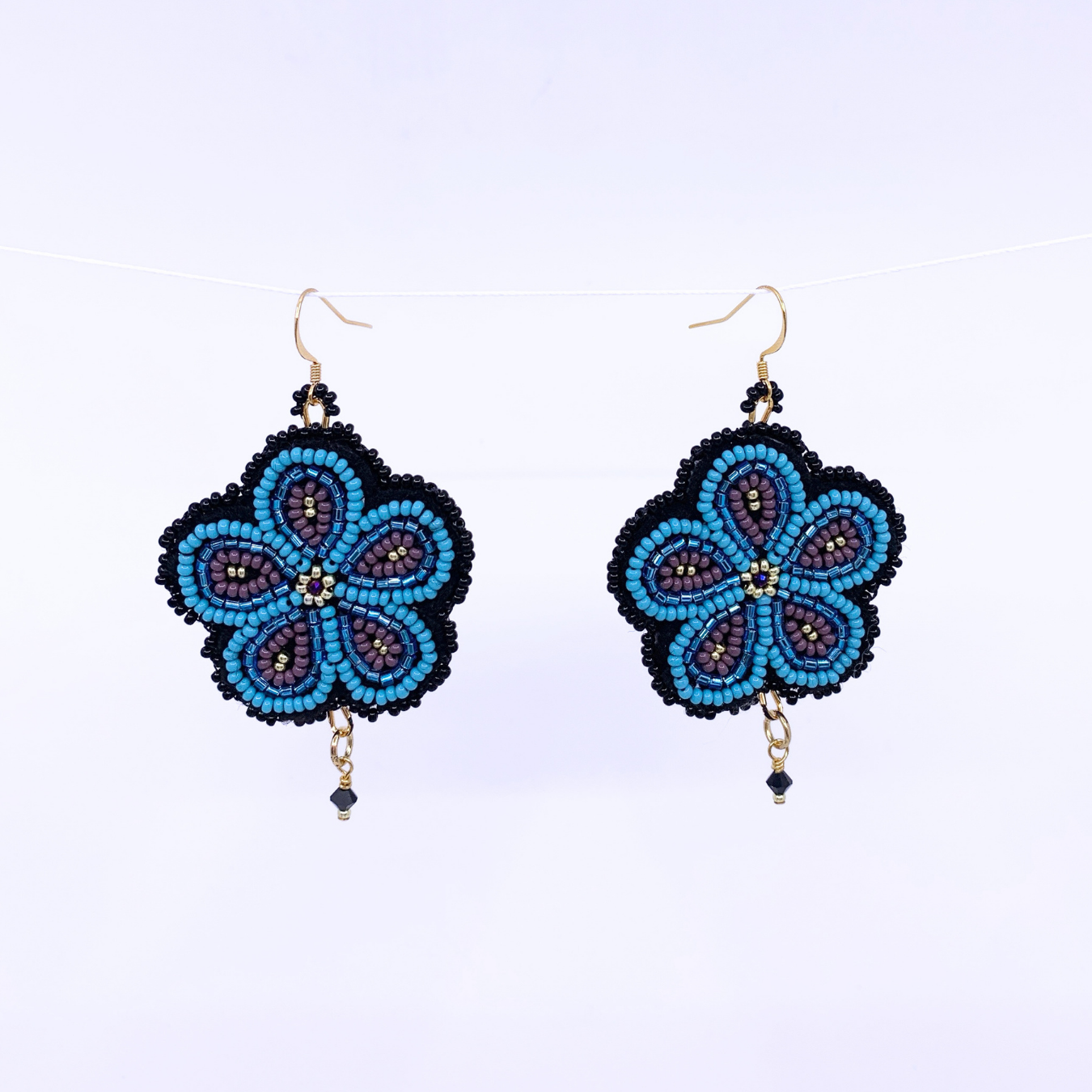 A pair of handmade beaded dangle earrings with a five-petal flower design in bright blue, purple, and black, hanging from gold-tone fishhook wires on a white background.