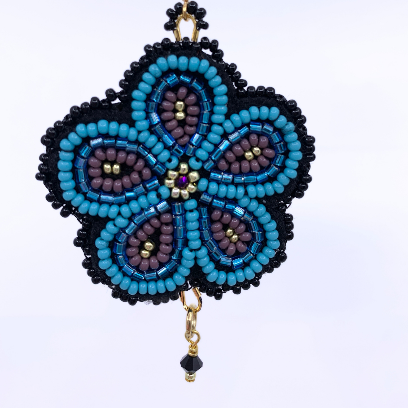 A handmade beaded flower ornament with a five-petal design, featuring turquoise blue, purple, and black seed beads with gold-tone metal accents and a dangling black bead at the bottom, set against a plain white background.