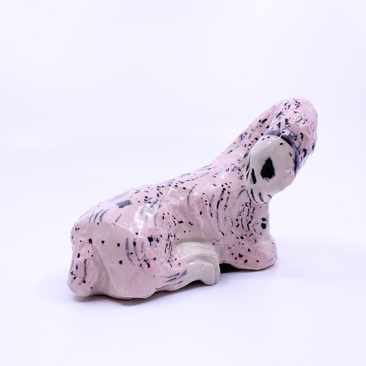 A small sculptural figurine of a kneeling animal, in a glossy pale pink glaze with black speckled and striped markings, photographed on a seamless white background.