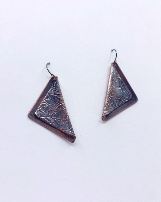 A pair of handmade geometric drop earrings with triangular shapes, featuring a textured, hammered metallic surface in a dark bronze and copper color, suspended from simple metal hooks on a plain white background.