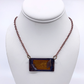 A rectangular pendant necklace displayed on a white mannequin bust. The pendant has a dark brown/black border with a textured, warm brown-orange centre, suspended from a dark copper-toned chain.