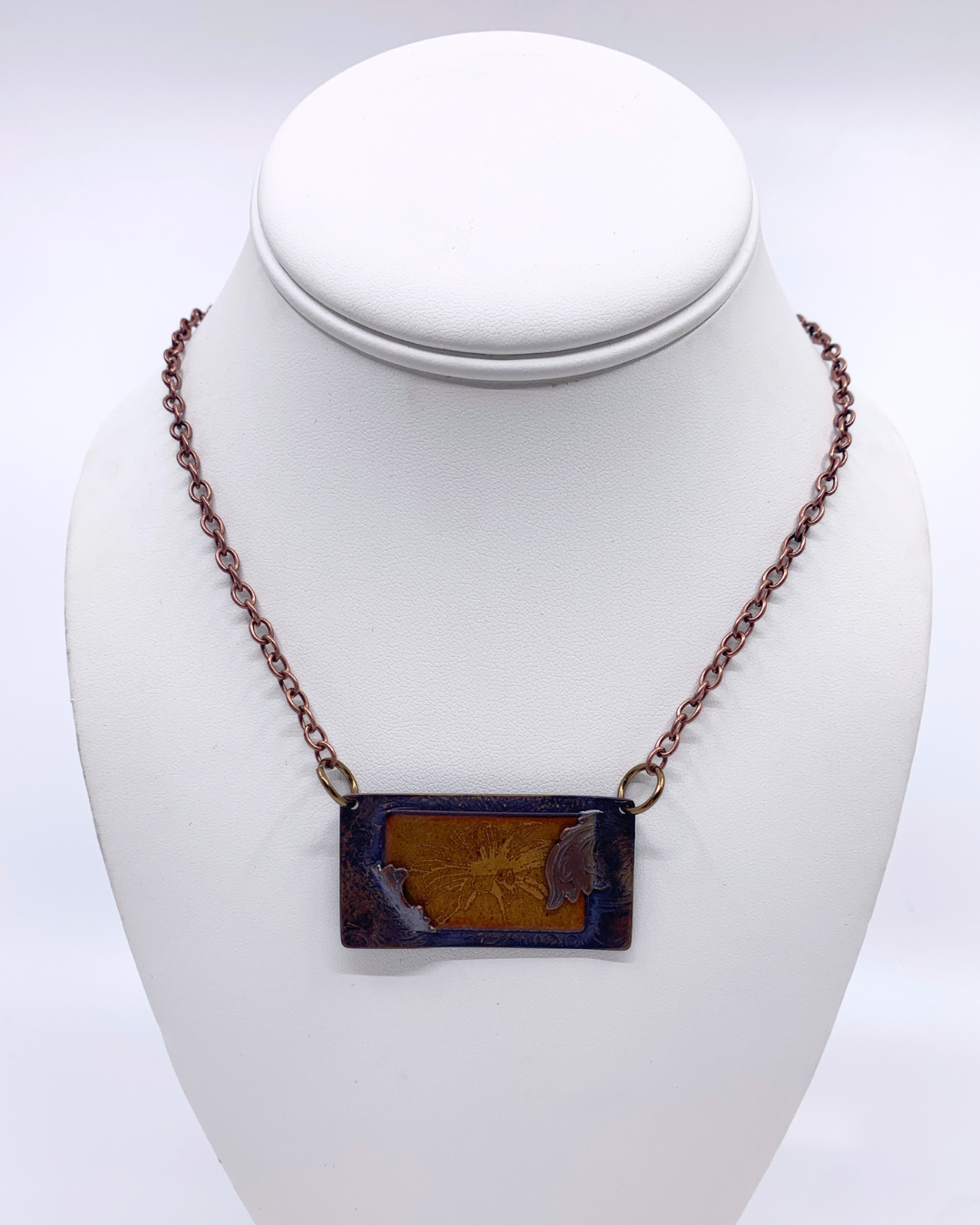 A rectangular pendant necklace displayed on a white mannequin bust. The pendant has a dark brown/black border with a textured, warm brown-orange centre, suspended from a dark copper-toned chain.