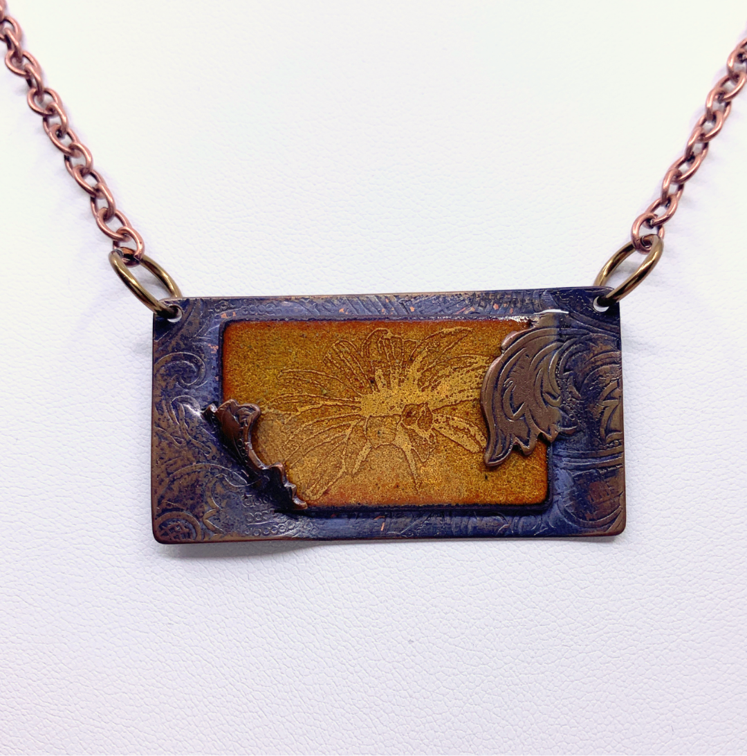 A rectangular pendant necklace with a copper-toned chain, featuring a central textured gold-toned panel with embossed floral details and a dark brown-grey border.