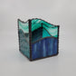 A square glass decorative box with a wavy, tiered design, featuring marbled teal, green, and blue glass panels framed by a dark, textured metallic border, set against a neutral light gray background.
