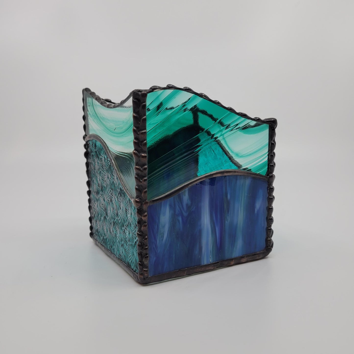 A square glass decorative box with a wavy, tiered design, featuring marbled teal, green, and blue glass panels framed by a dark, textured metallic border, set against a neutral light gray background.