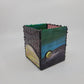 A small decorative box with green, blue, yellow, and purple hues, framed by a dark metallic border with rivet details.