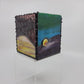 A small decorative box with green, blue, yellow, and purple hues, framed by a dark metallic border with rivet details.