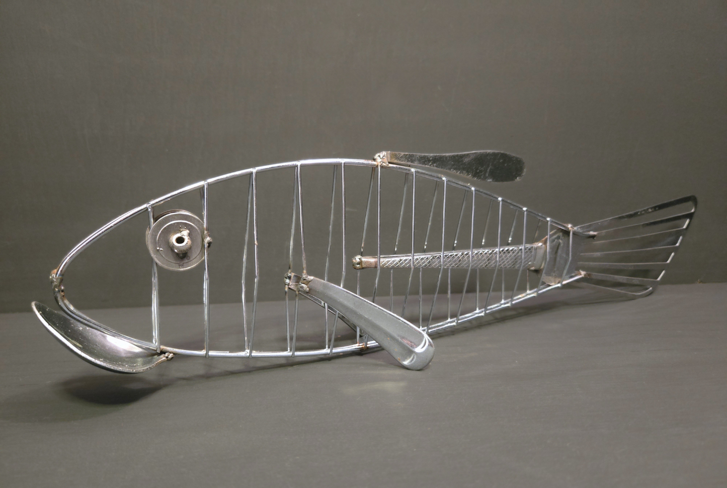 Metal fish made from repurposed cutlery, silver in colour, on grey background
