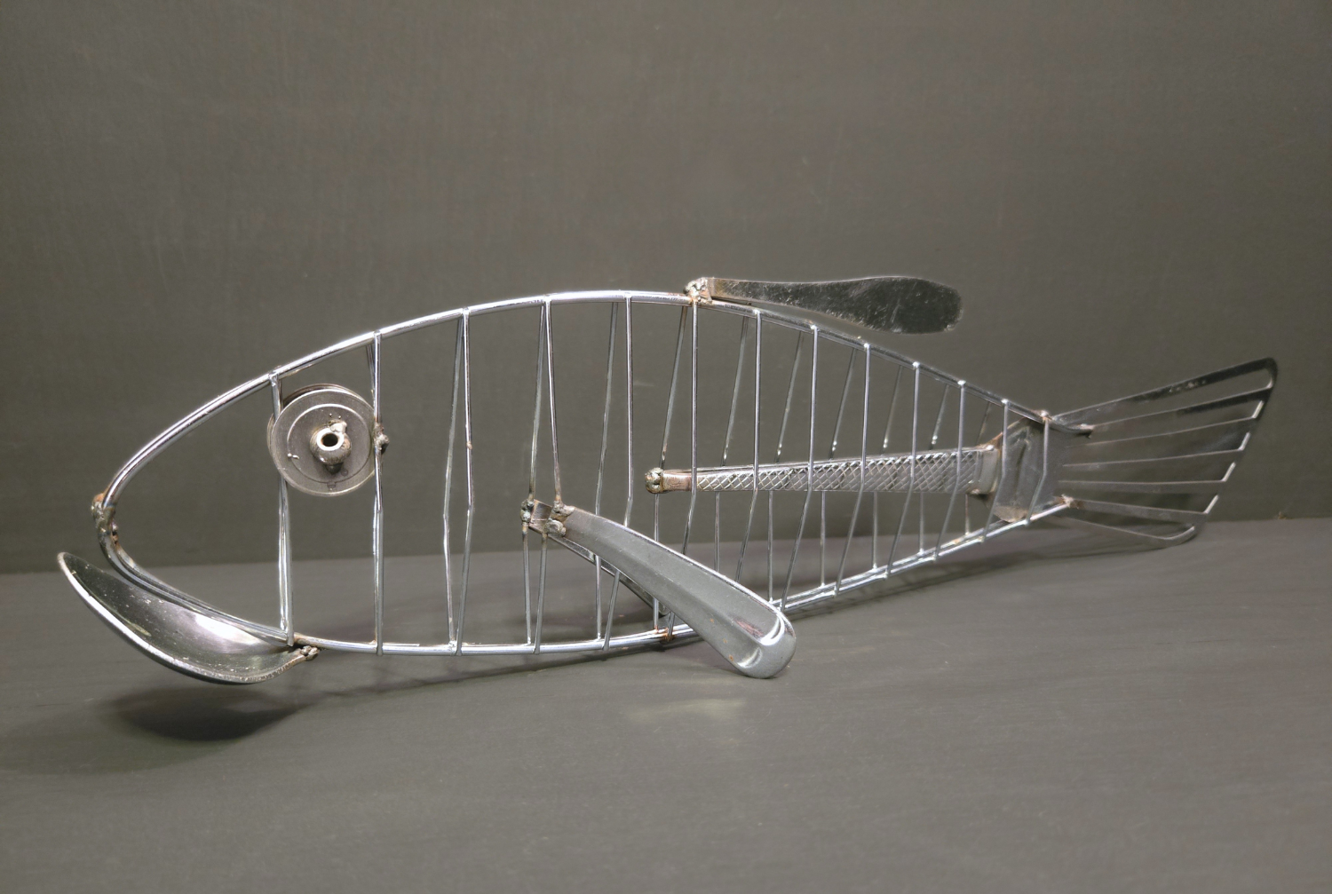 Metal fish made from repurposed cutlery, silver in colour, on grey background