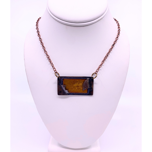 A rectangular pendant necklace with a dark amber-brown marbled design, hanging on a copper-toned chain from a white mannequin bust.
