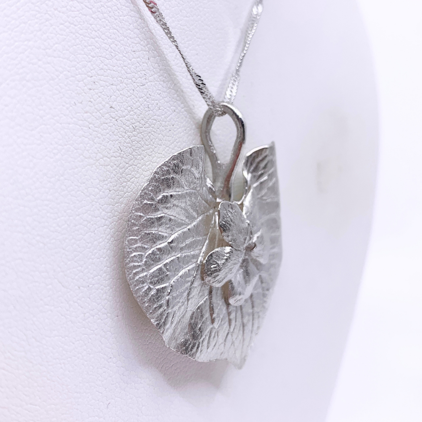 A silver pendant necklace featuring a textured, organic leaf-like shape with a small raised floral detail, displayed on a white curved jewelry bust.