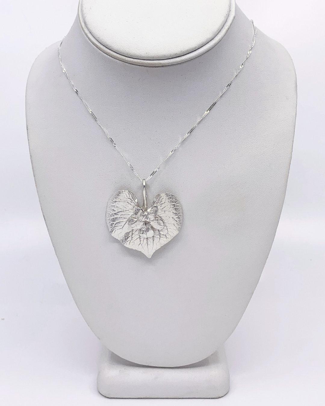 A silver pendant necklace featuring a textured, leaf-like heart design with a small flower in the center, displayed on a white jewelry bust.