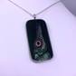 A rectangular silver pendant necklace featuring a dark, textured design with a stylized fish eye motif in pink, green, and black, suspended from a silver chain on a white background.