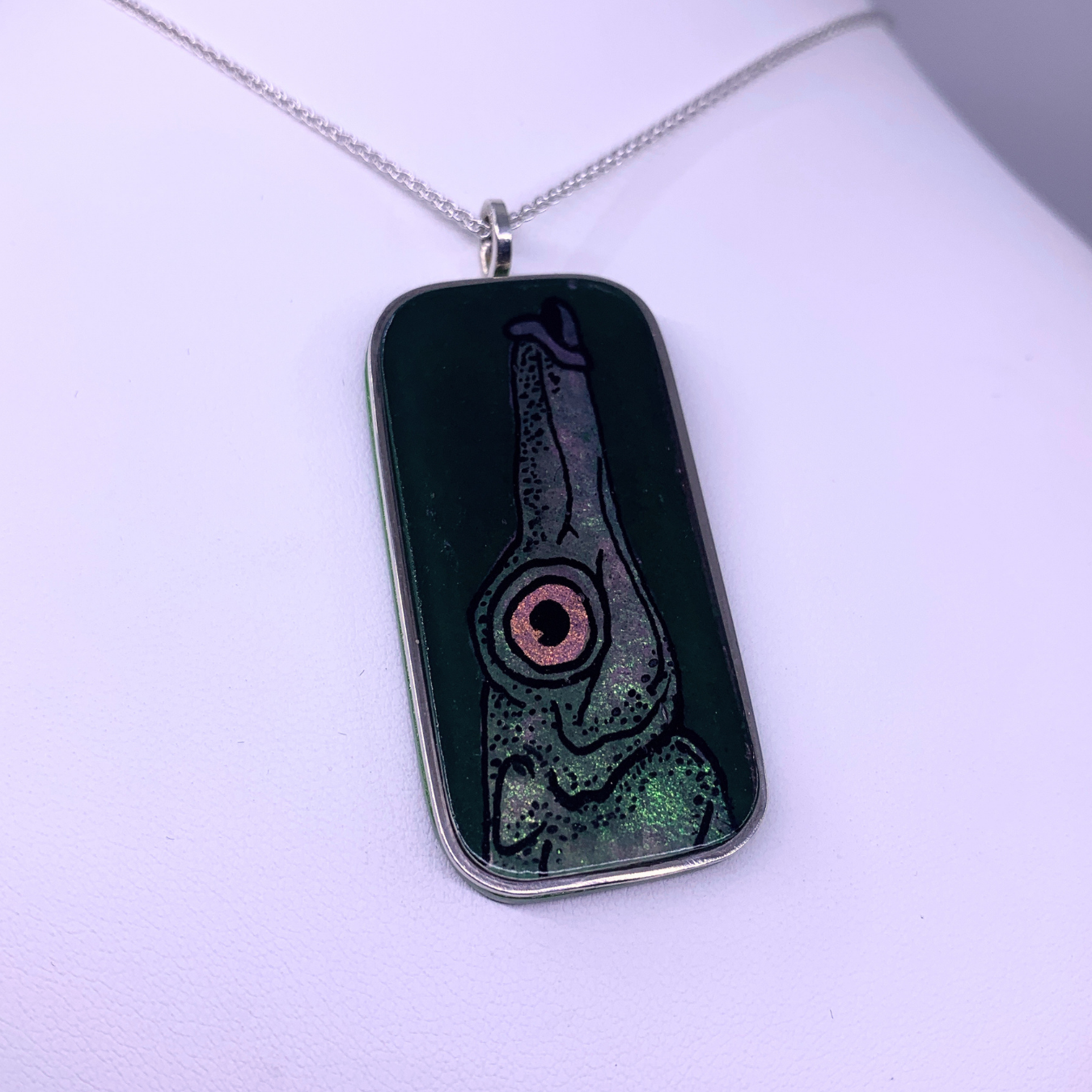 A rectangular silver pendant necklace featuring a dark, textured design with a stylized fish eye motif in pink, green, and black, suspended from a silver chain on a white background.