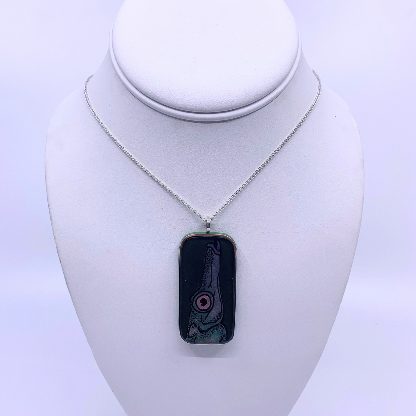A silver chain necklace with a rectangular pendant featuring a black background and a stylized dish design in pink, red, and green, displayed on a white neck mannequin.