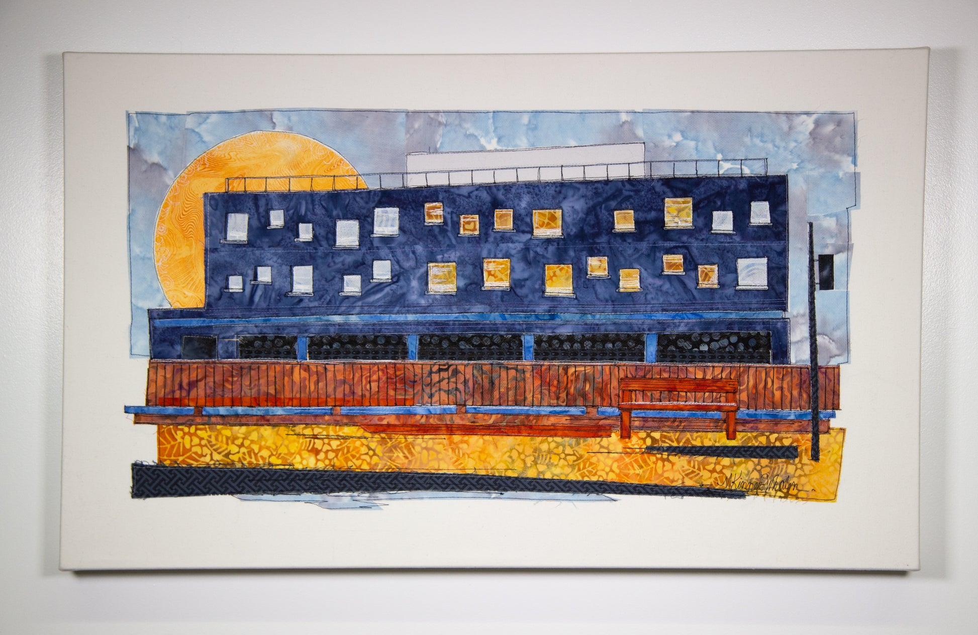 Quilted thread painting of a blue building with many windows. A fence is in front with a bench. 